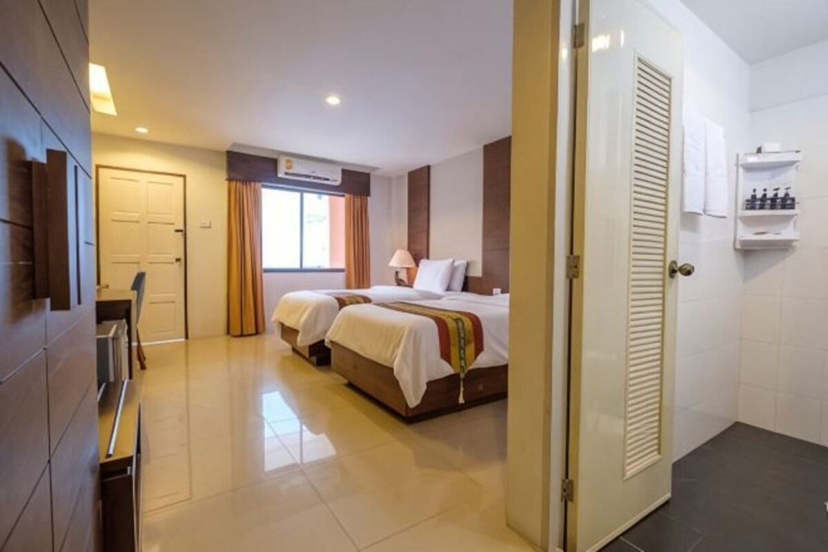 A hotel room with two beds, a desk, a chair, a window with curtains, an air conditioning unit, and a door.