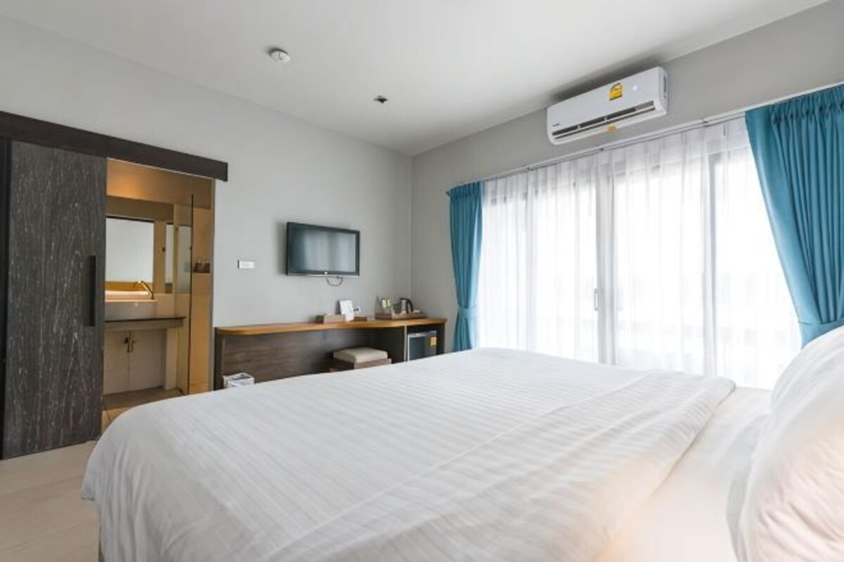 A hotel room with a bed, a desk, a mirror, a television, and an air conditioning unit.