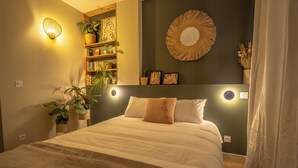 A neatly made bed with a headboard, a bedside shelf with books and plants, and a wall-mounted mirror. - Cocon Urban Jungle - Studio Rénové Dans le Centre Ancien (Montélimar)