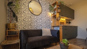 A living room with a dark sofa, a wooden coffee table, and a kitchen area with a sink and cabinets. - Cocon Urban Jungle - Studio Rénové Dans le Centre Ancien (Montélimar)