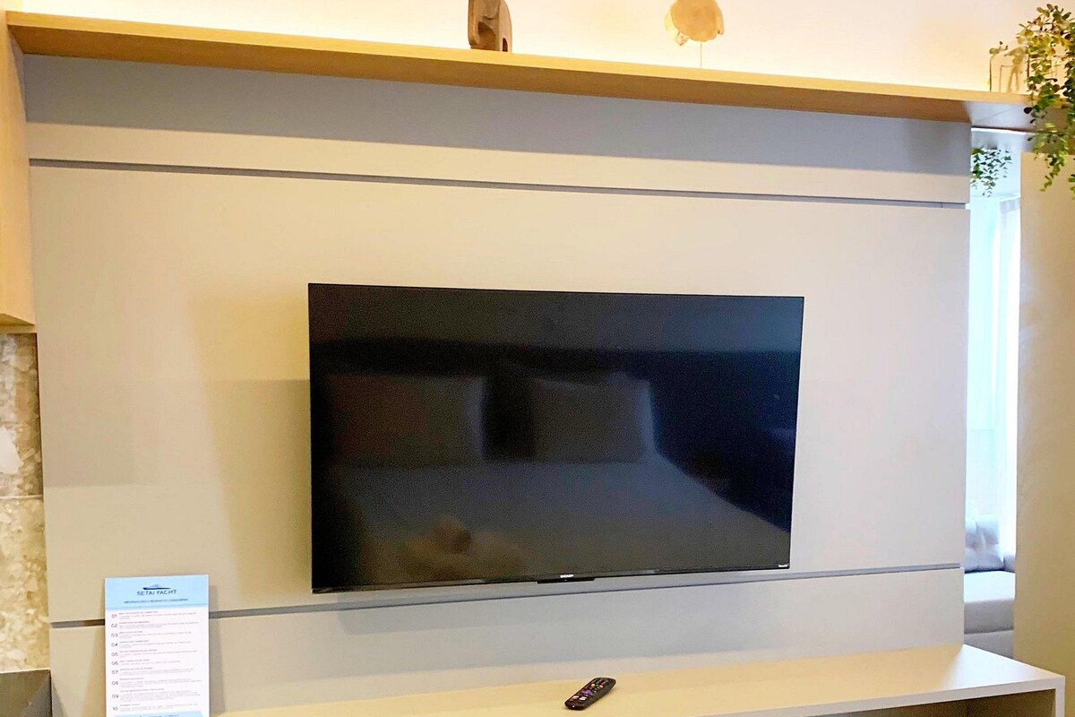 Une chambre d’hôtel moderne équipée d’un téléviseur à écran plat fixé au mur, d’une petite télécommande, d’un coin salon avec un banc et d’une serviette soigneusement pliée sur le lit.