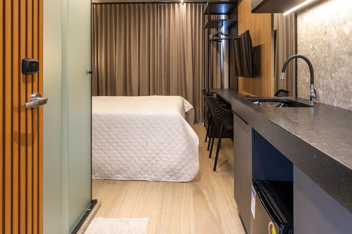 A compact hotel room with a kitchenette, a bed, a seating area, and a wooden door.
