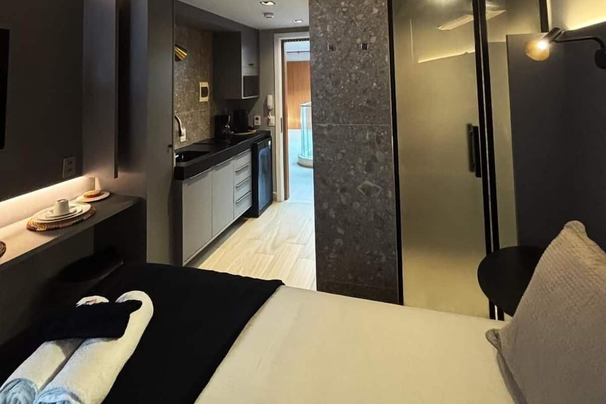 A modern hotel room with a bed, a desk, a bathroom, and a kitchen area.