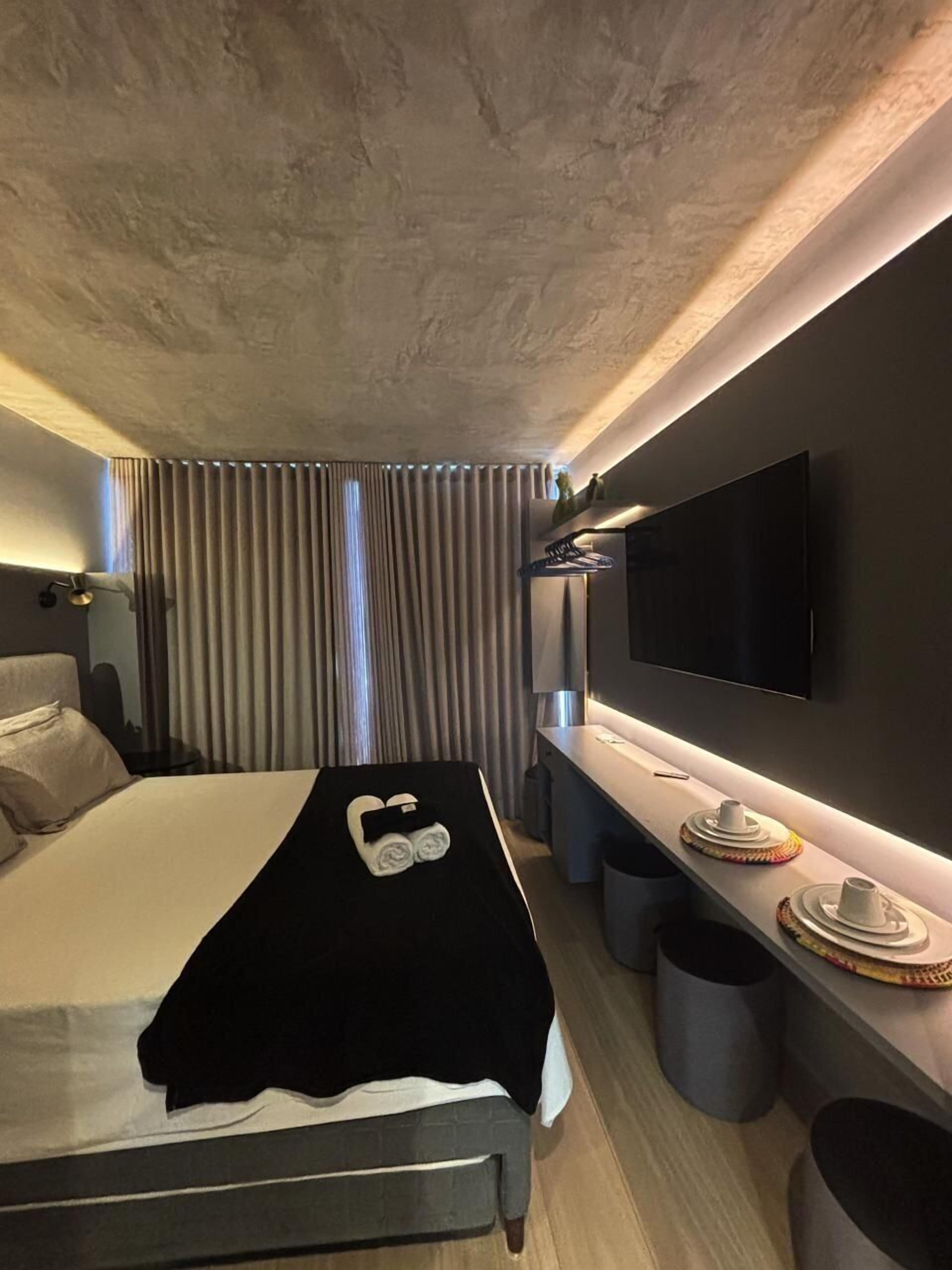 Ein modernes Hotelzimmer mit Bett, Wandfernseher, Schreibtisch mit Stuhl und einem Vorhangfenster.