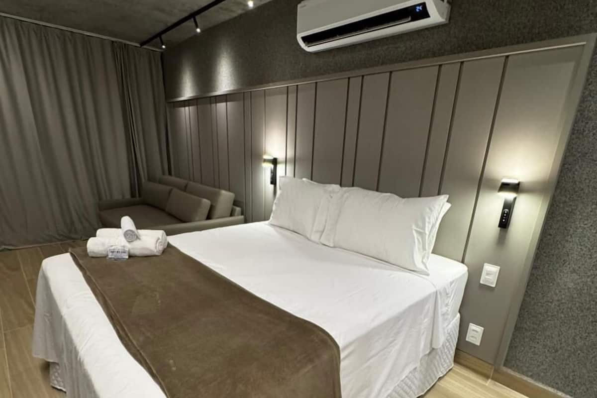 A modern hotel room with a large bed, a sofa, an air conditioning unit, and a bedside table.