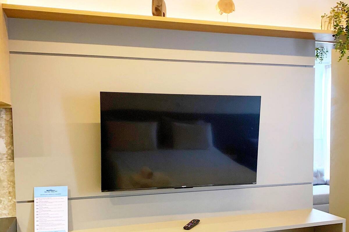 A modern hotel room with a flat-screen TV mounted on a wall, a small remote control, a seating area with a bench, and a neatly folded towel on the bed.
