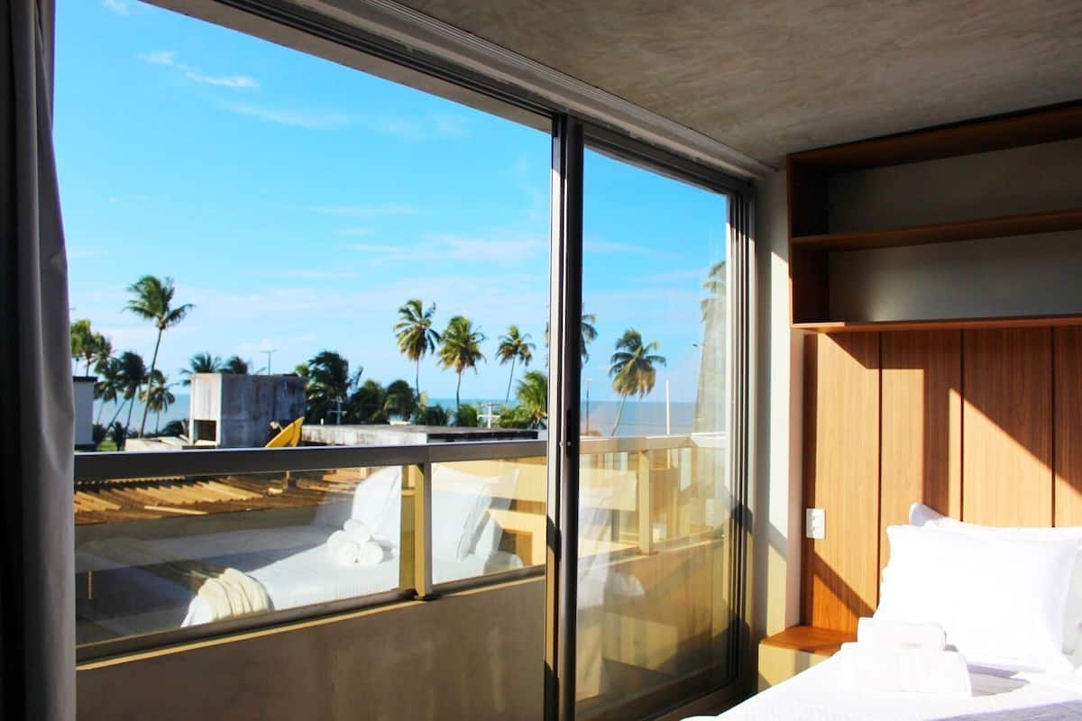 A hotel room with a large sliding glass door, a bed with white linens, a wooden headboard, and a view of palm trees and a beach.