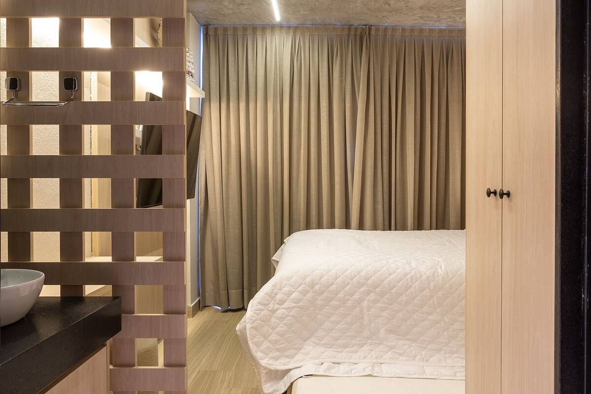 A bedroom with a bed, a wooden wardrobe, a bathroom with a sink, and a partition with a light fixture.
