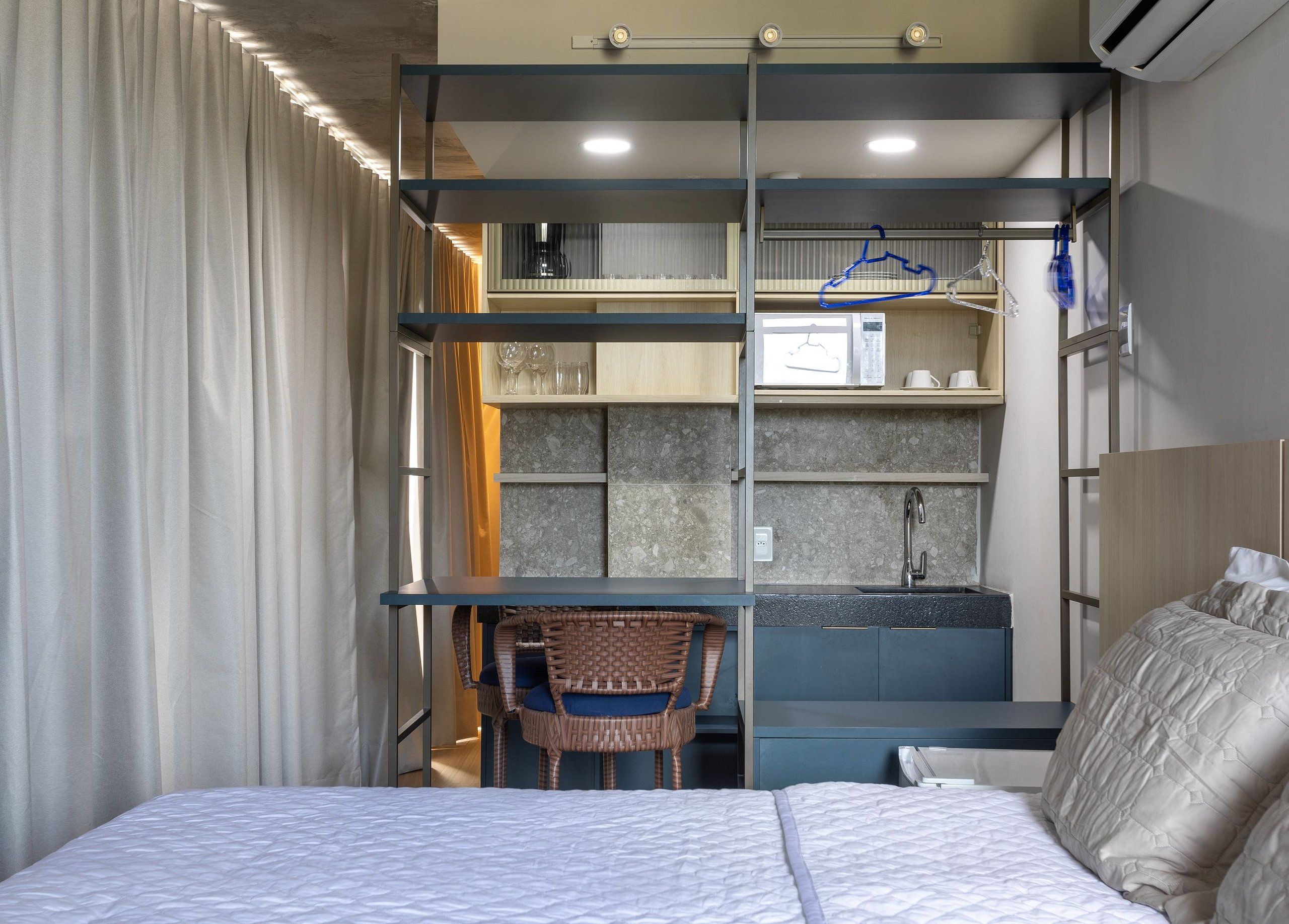 Kompakti hotellihuone, jossa on sänky, pieni työpöytä, tuoli ja pieni keittiönurkkaus.