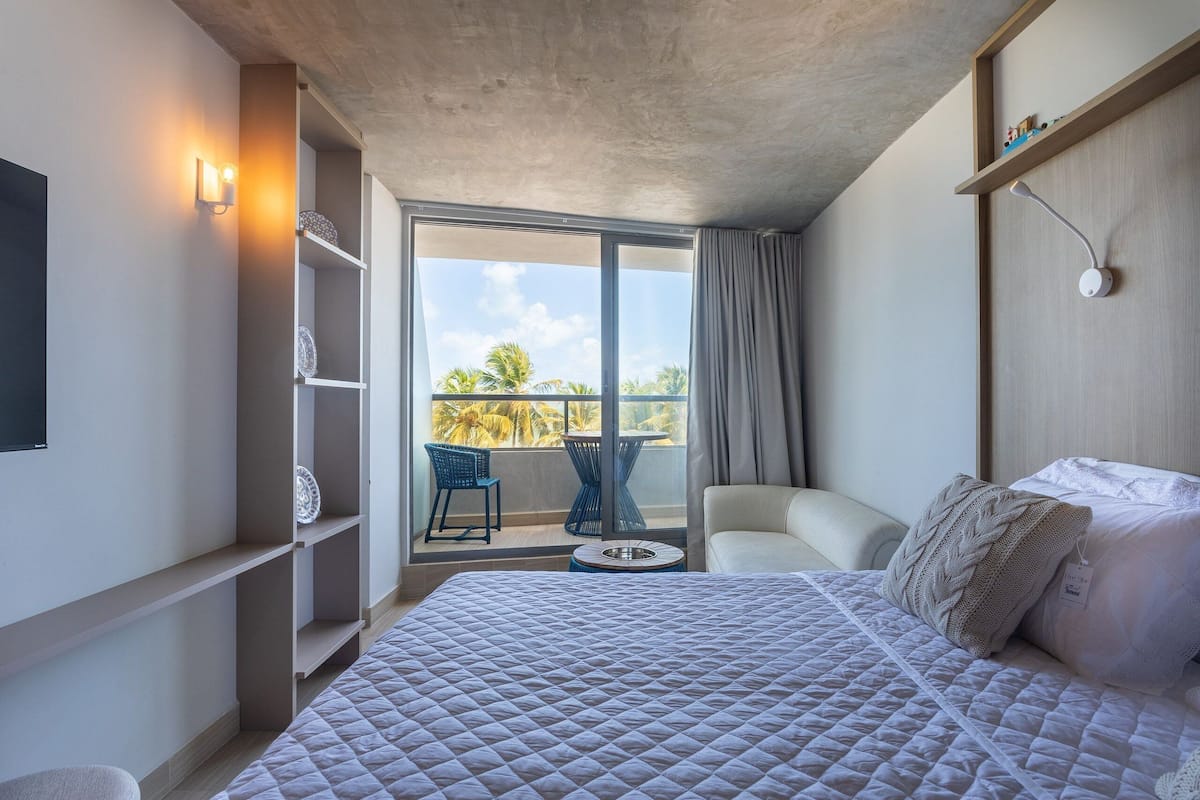 A hotel room with a bed, a chair, a small table, a balcony with a view, and a shelf with decorative items.