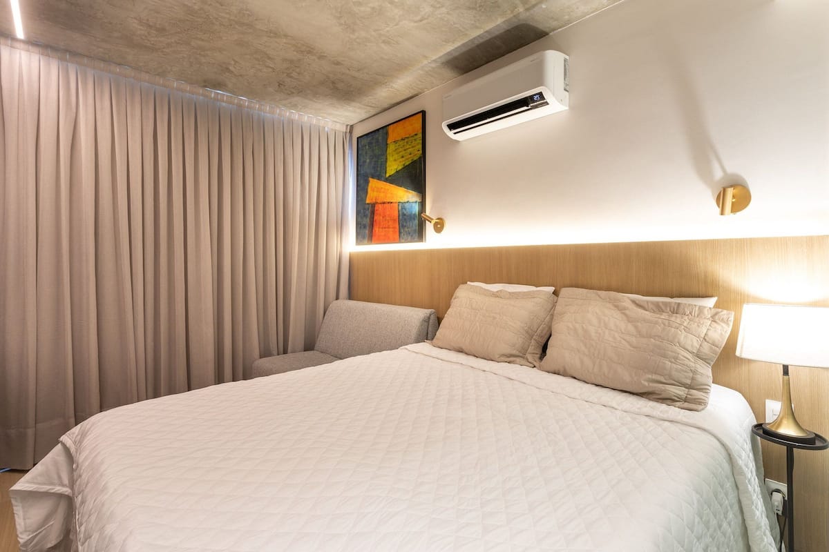 A hotel room with a bed, a bedside lamp, an air conditioning unit, and a painting on the wall.
