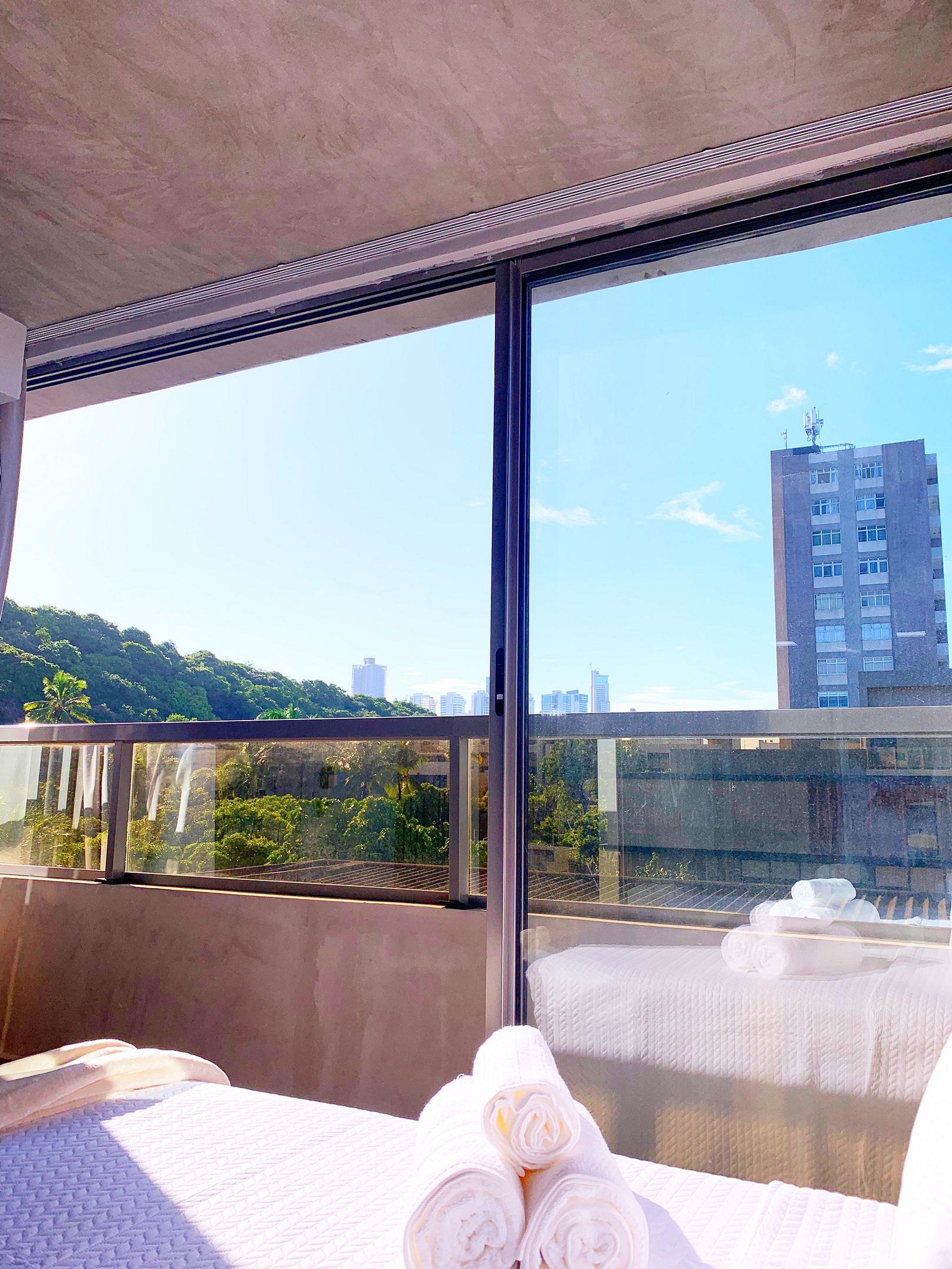 Hotellihuone, jossa on suuri ikkuna, valkoisilla lakanoiden peitetty sänky sekä näkymä kaupunkimaisemaan ja vihreälle alueelle.