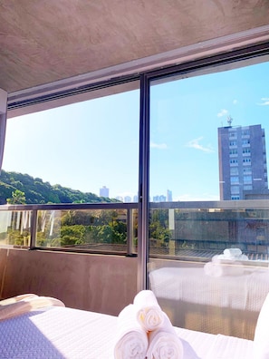 Une chambre d’hôtel dotée d’une grande fenêtre, d’un lit avec du linge de maison blanc, et offrant une vue sur le paysage urbain et la verdure.