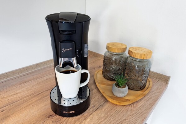 Une machine à café avec une tasse posée sur un plateau, deux bocaux avec des couvercles et une petite plante sur une table en bois.