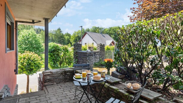 Un patio con mesa lista para el desayuno, un muro de piedra y vistas a una casa y árboles.