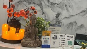 Una statua raffigurante una figura seduta, un vaso con bacche rosse e un candelabro con candele accese. Sul tavolo sono presenti cartelli informativi e un codice QR.