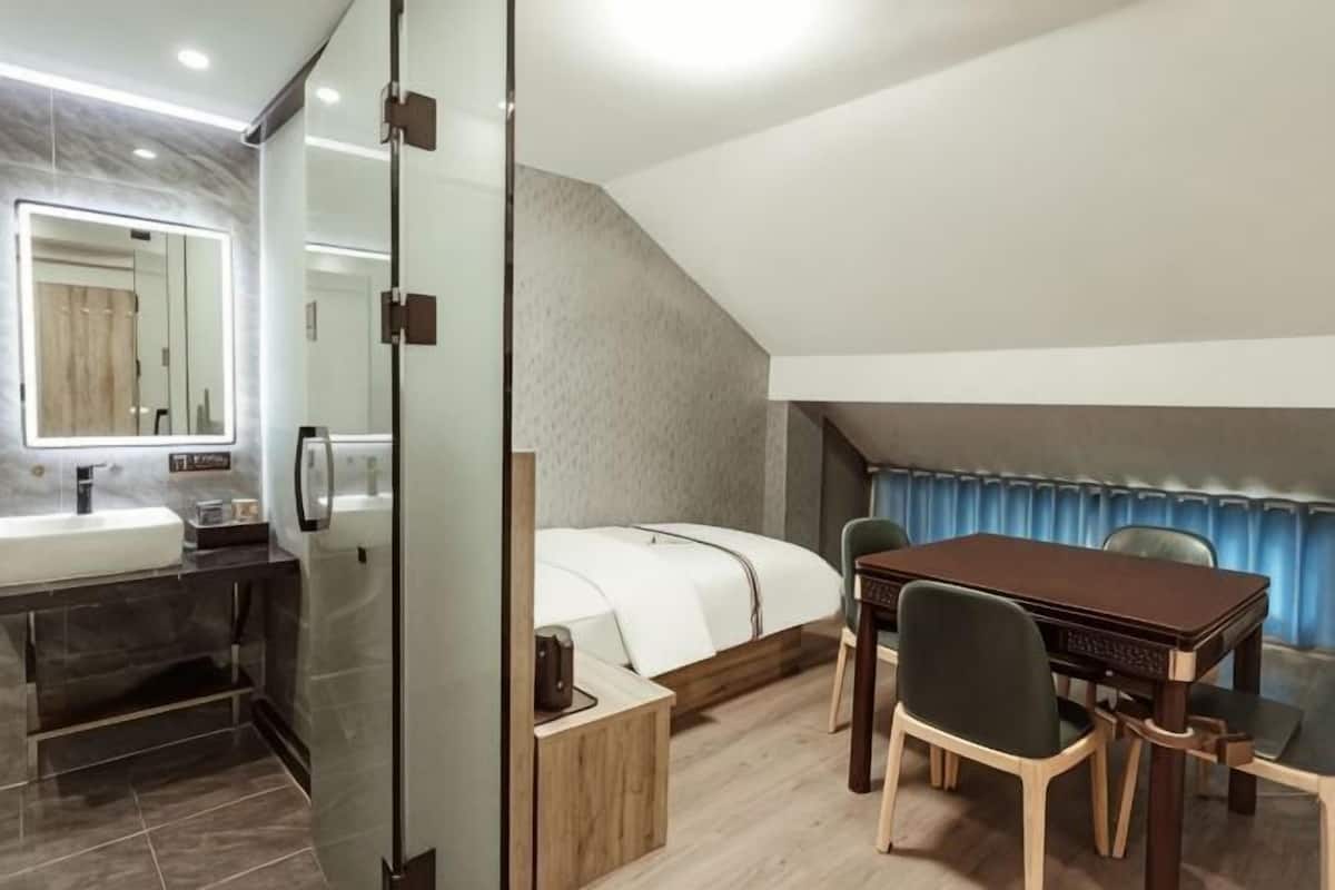 A modern bedroom with a glass partition, a bed, a wooden table, and a bathroom with a sink and mirror.