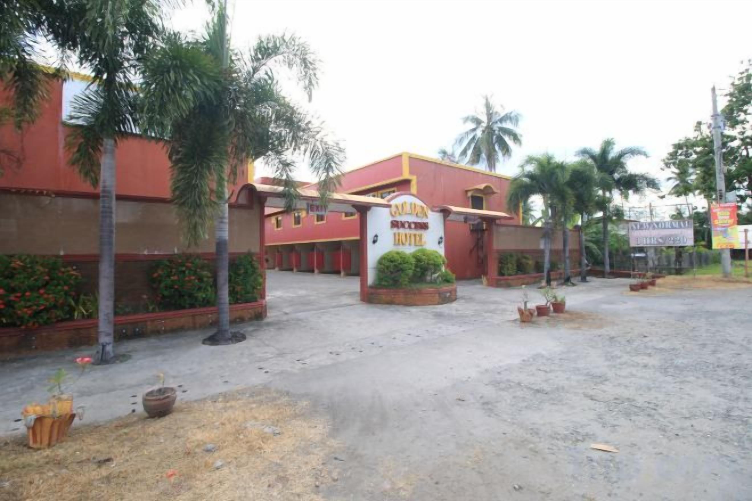 Entrada de hotel con un cartel, palmeras y una zona pavimentada.