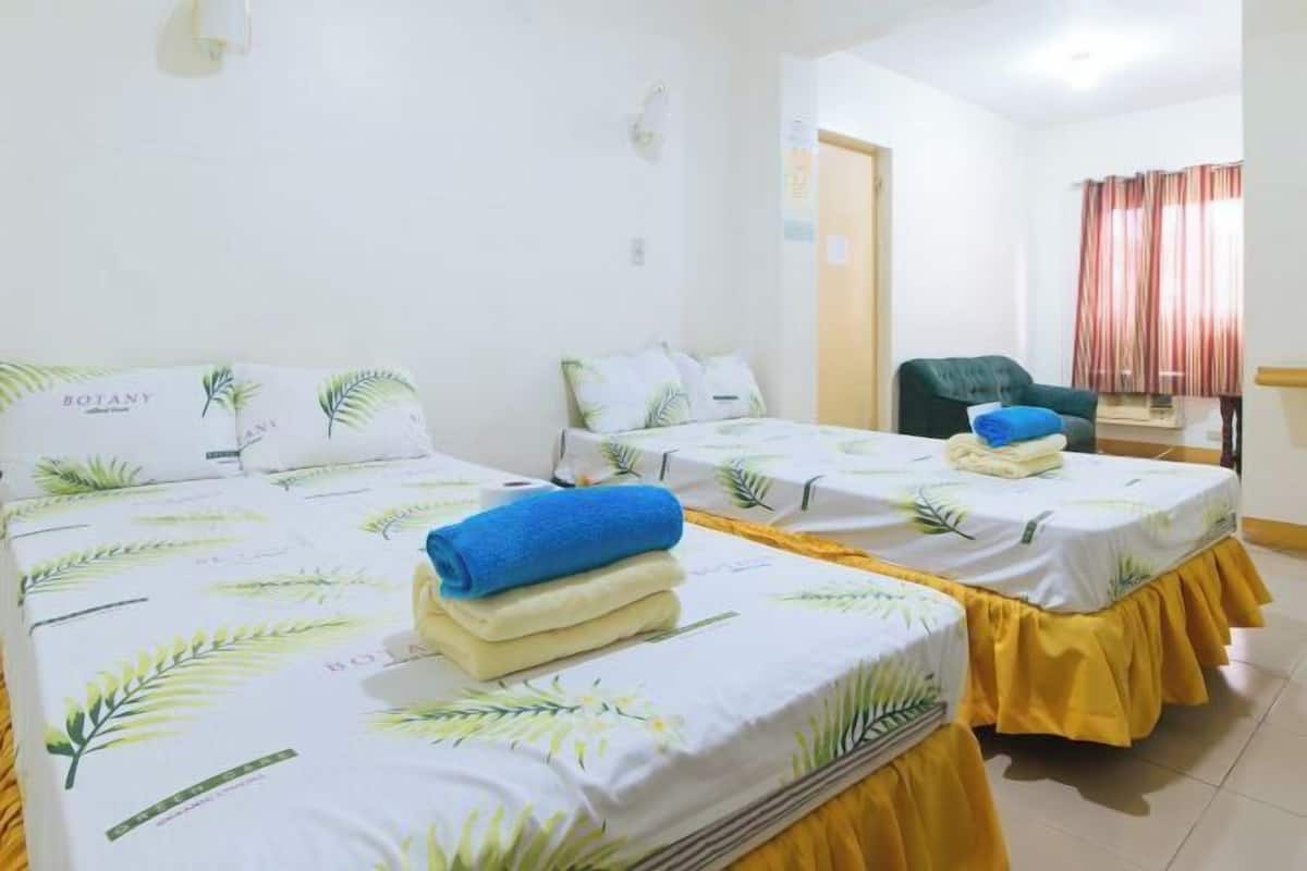 A hotel room with two beds, each with a white bedspread featuring palm leaf patterns and a yellow skirt. There is a chair and a window with curtains.