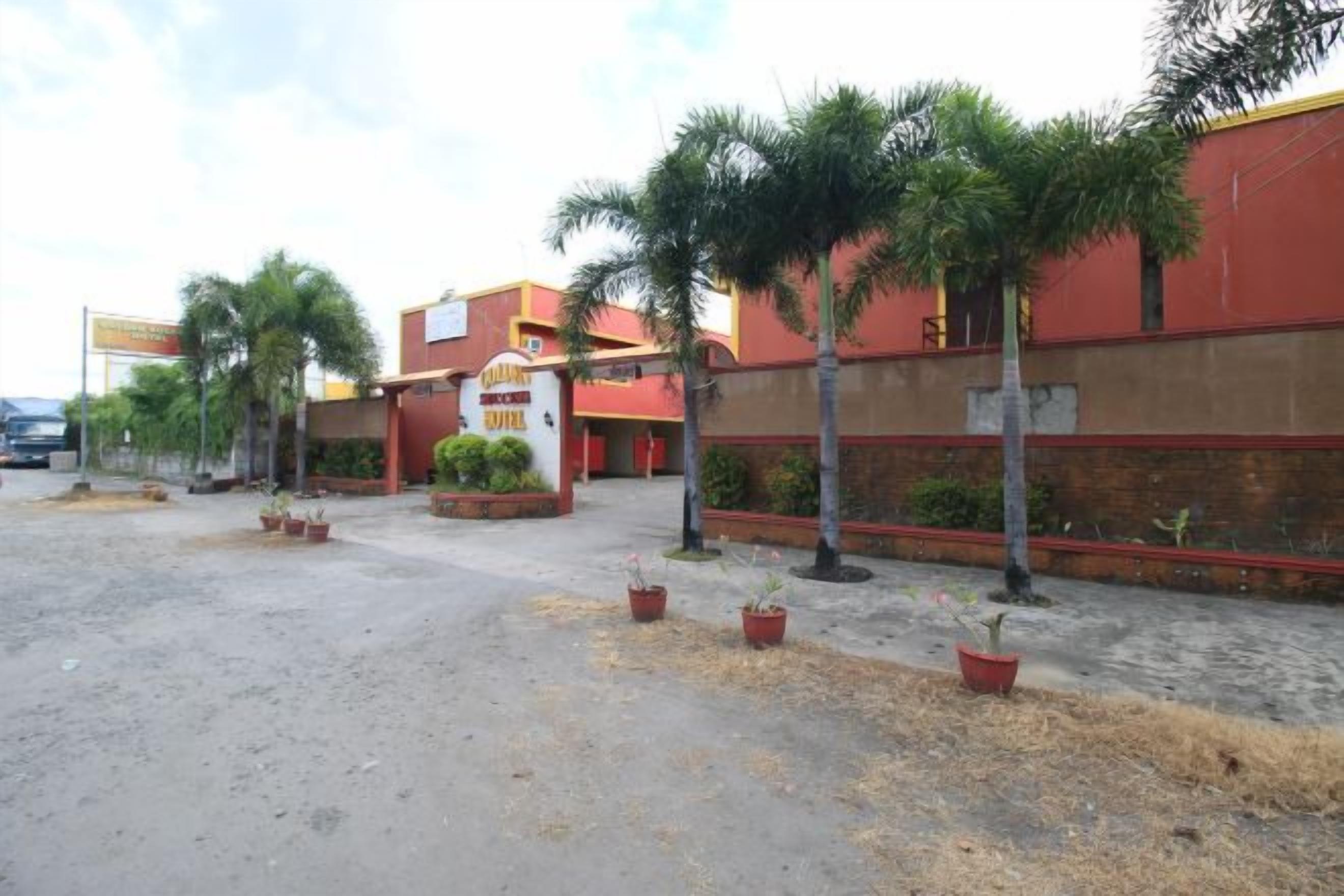 Fachada roja de un hotel, palmeras y un camino pavimentado.