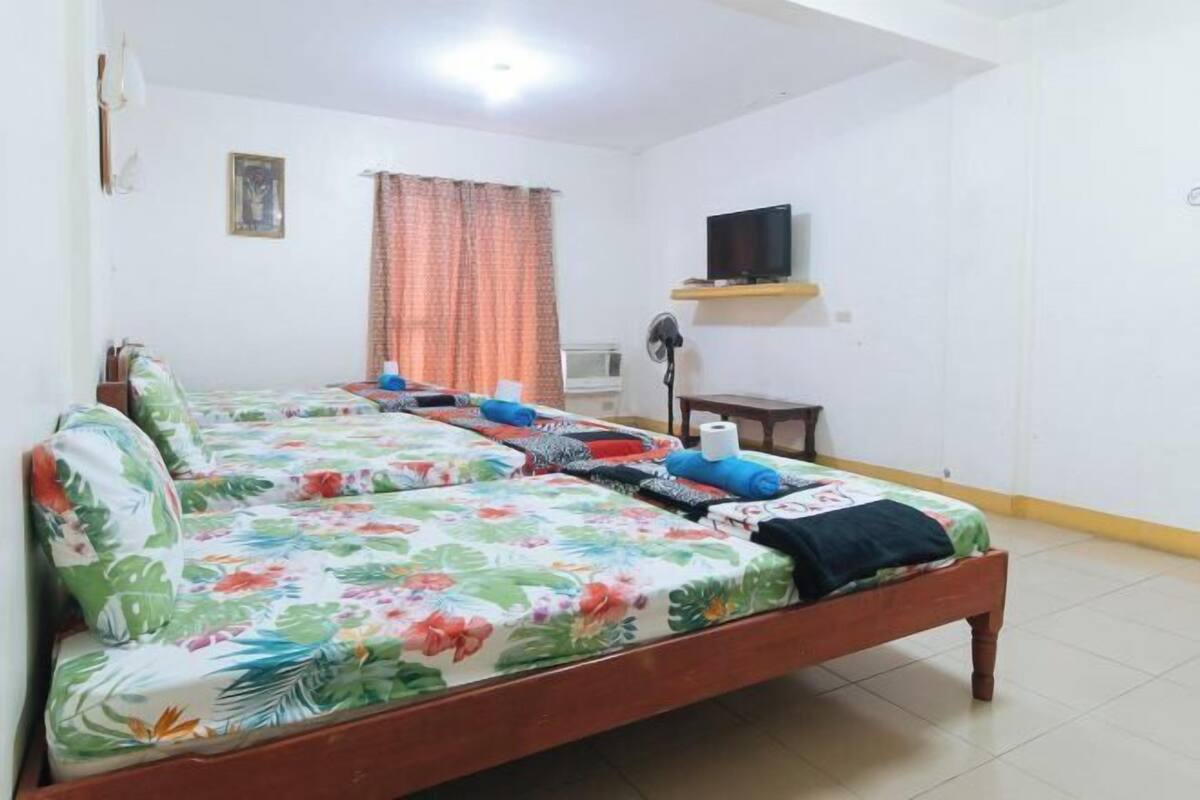 A room with a large bed, a flat-screen TV mounted on the wall, a ceiling fan, and a window with curtains.