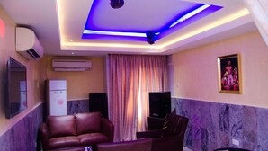 Ein Zimmer mit blauer LED-Beleuchtung an der Decke, einem großen Fenster mit Vorhängen, einem Flachbildfernseher, einem Kühlschrank und einem Sofa.