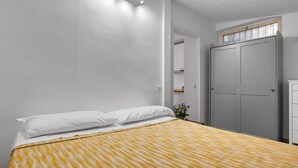 A bedroom with a bed, white pillows, a yellow-striped bedspread, a nightstand with a vase of flowers, a wardrobe, and a window with blinds. - Colosseum Street Haven (ROMA)
