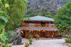 A rustic building with a thatched roof, a balcony, and a garden area with potted plants and a rock formation. - The Goat Boutique Resort (Tay Hoa Lu)