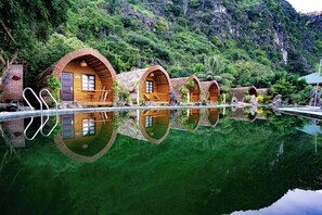 Une rangée de charmantes chaumières aux toits de chaume, dotées de balcons en bois, une piscine et une végétation luxuriante en arrière-plan.