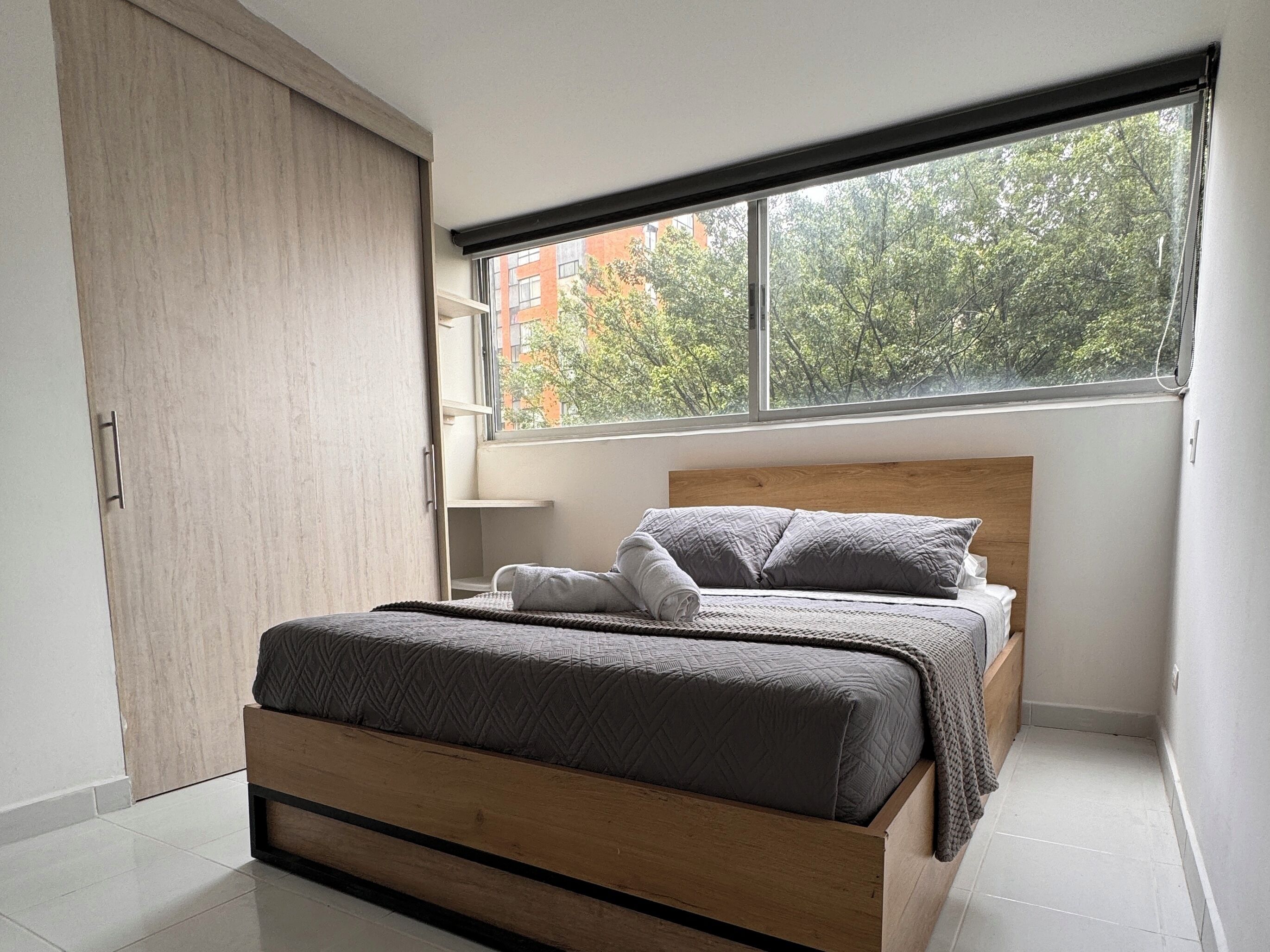 Un dormitorio moderno con cama de madera, un ventanal con vistas a árboles y edificios, y un armario con puerta corredera.