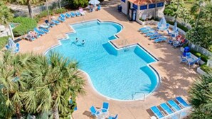 Ein Pool mit ungewöhnlicher Form, umgeben von Liegestühlen und Sonnenschirmen, inmitten einer angelegten Grünfläche mit Palmen.
