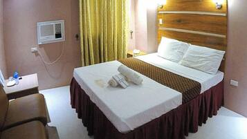 A hotel room with a bed, a desk, a chair, a nightstand, a lamp, and a window with curtains.