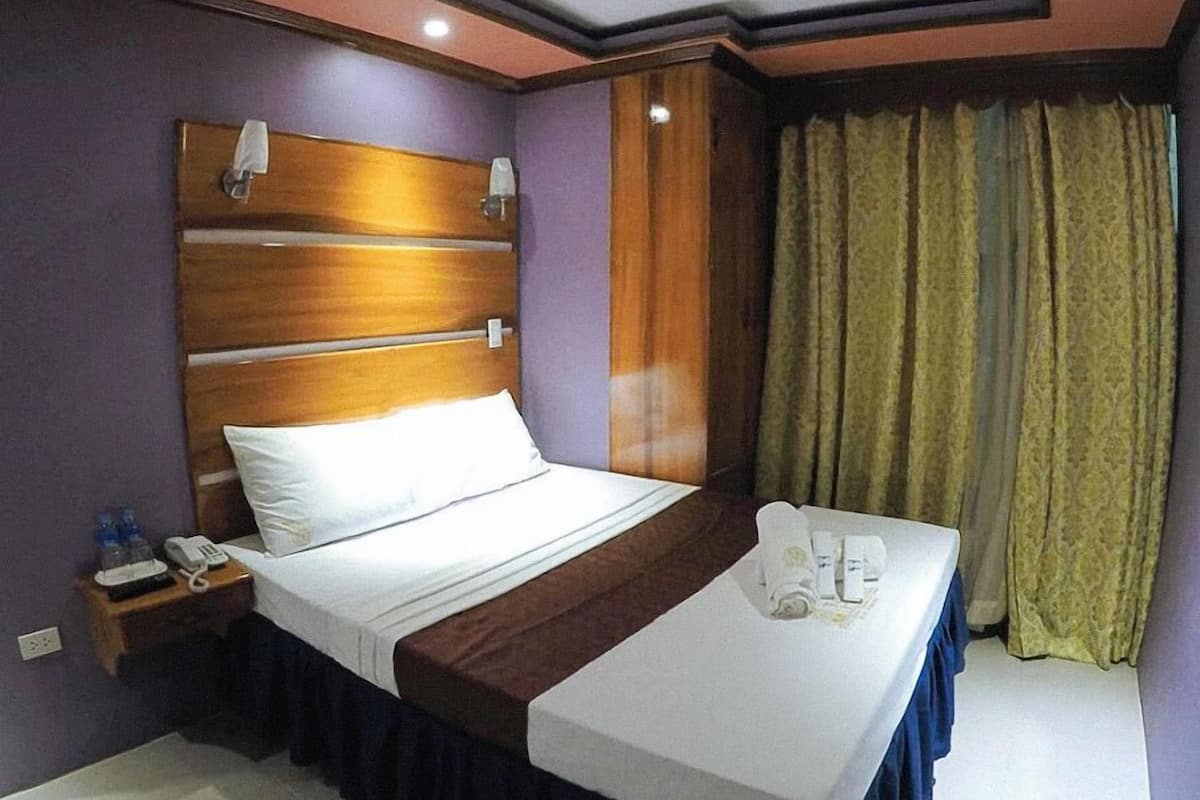 A hotel room with a wooden headboard, a neatly made bed, a bedside table with a phone and water bottles, and a window with curtains.