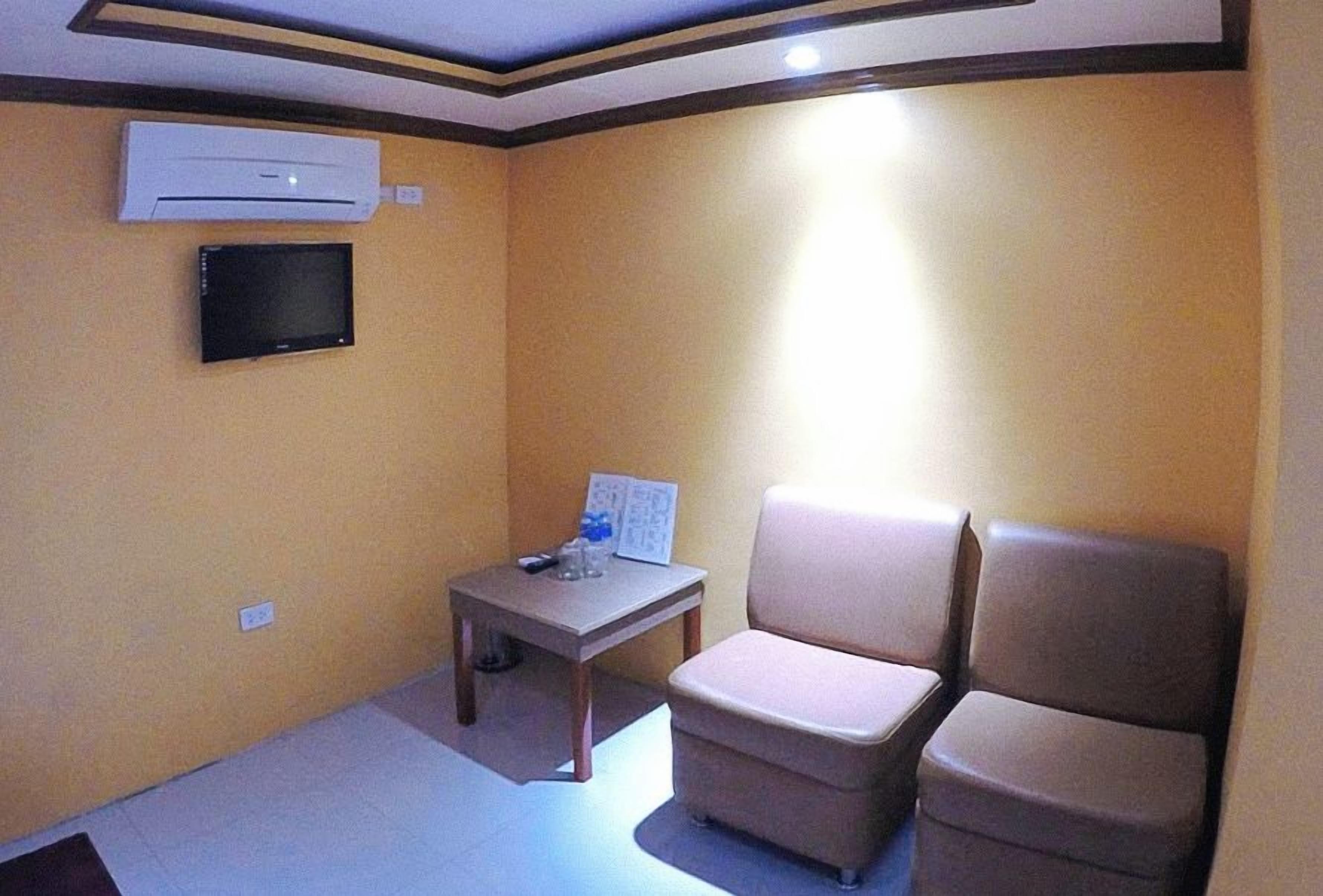 A small room with a beige wall, a flat-screen TV mounted on the wall, a small table with a remote control, and two beige chairs.