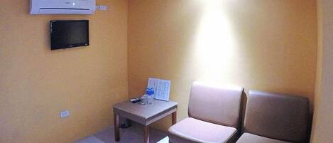 A small room with a beige wall, a flat-screen TV mounted on the wall, a small table with a remote control, and two beige chairs.