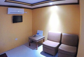 Una habitación pequeña con una pared color beige, un televisor de pantalla plana montado en la pared, una mesita con un control remoto y dos sillas color beige.