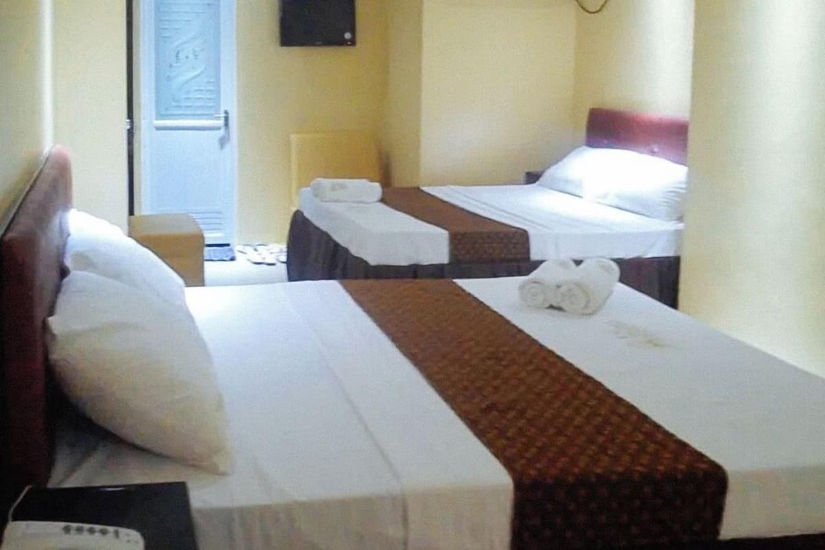 A hotel room with two beds, each with white linens and brown headboards. There is a small table with a lamp and a box on it.