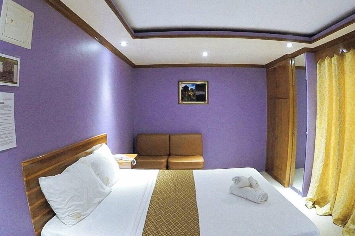 A hotel room with a bed, a brown sofa, a small table, and a window with curtains.