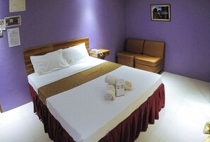 Habitación de hotel con cama, mesita de noche, sillón marrón y una pequeña mesa con un teléfono y papeles.