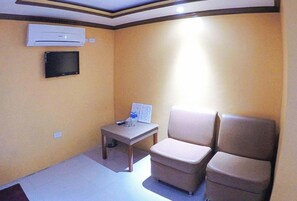 Een kleine kamer met een beige bank, een tafeltje met een afstandsbediening en een airconditioner aan de muur.