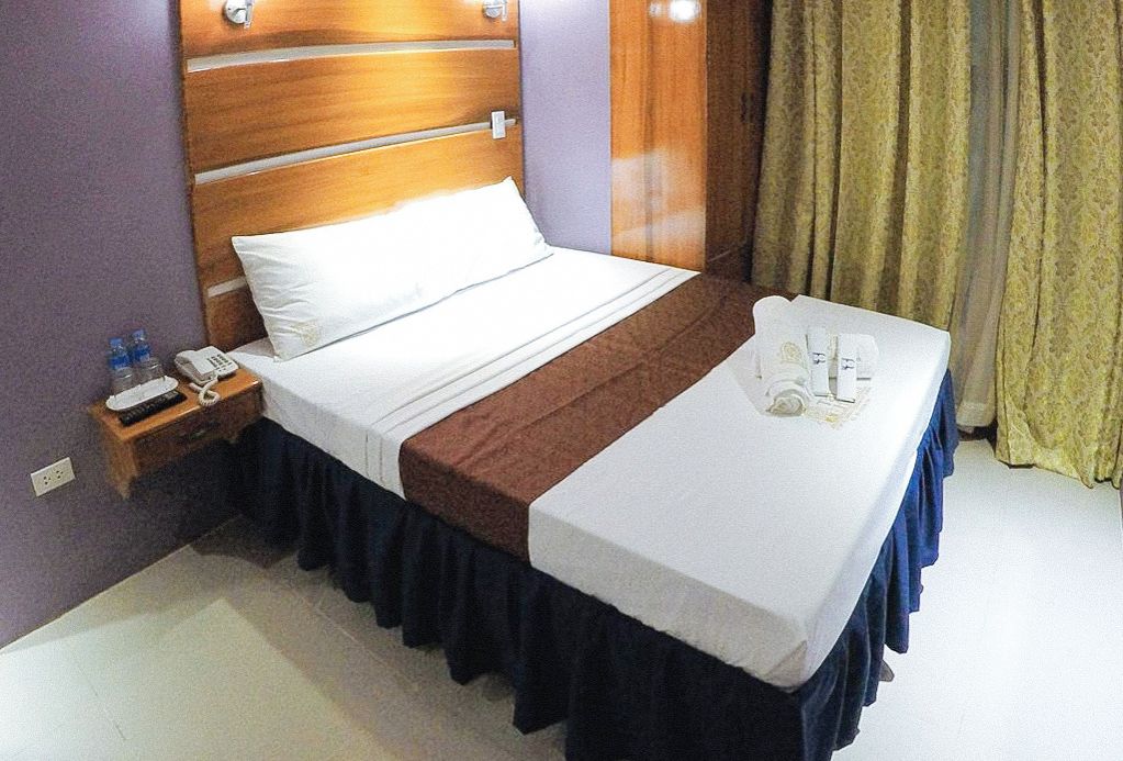 A hotel room with a wooden headboard, a bed with white and brown linens, a bedside table with a phone and water bottles, and a window with curtains.