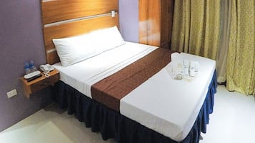 A hotel room with a wooden headboard, a bed with white and brown linens, a bedside table with a phone and water bottles, and a window with curtains.
