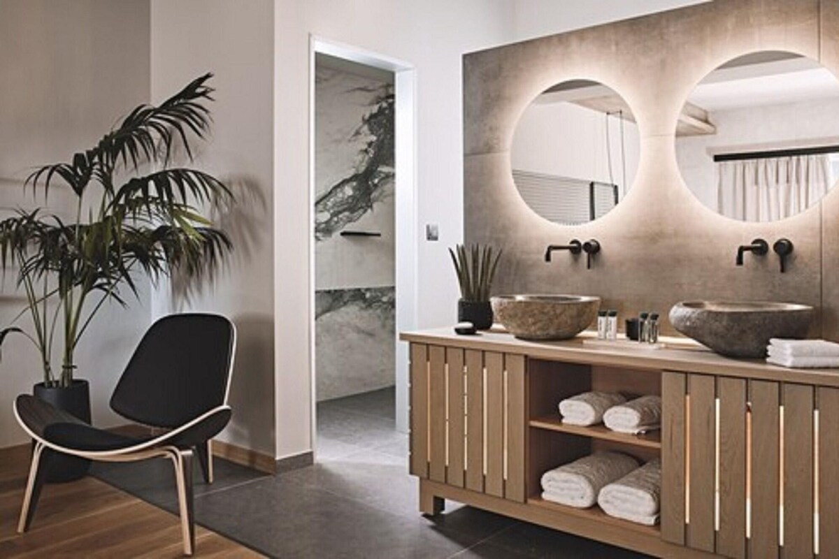 Un bagno moderno con un mobile vanitario in legno, due specchi rotondi, una pianta in vaso e una sedia nera.