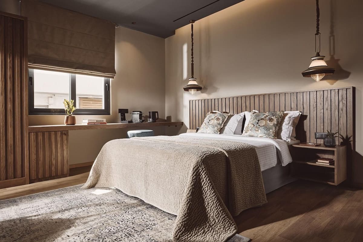 Ein Schlafzimmer mit hölzernem Kopfteil, einem großen Bett mit vielen Kissen, Nachttischen, einem Fenster mit Jalousien und einer Hängeleuchte.