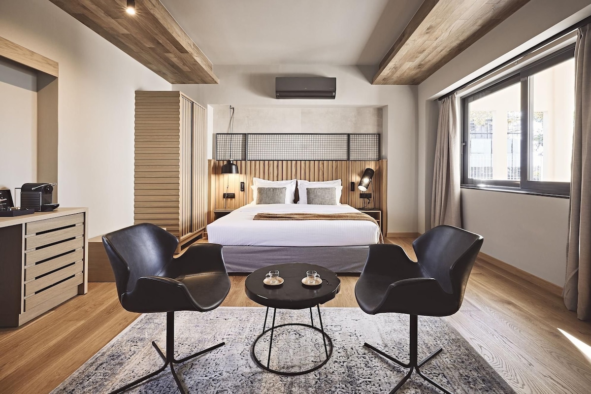 A modern hotel room with a large bed, two black chairs, a small table, a wooden desk, and a large window with curtains.