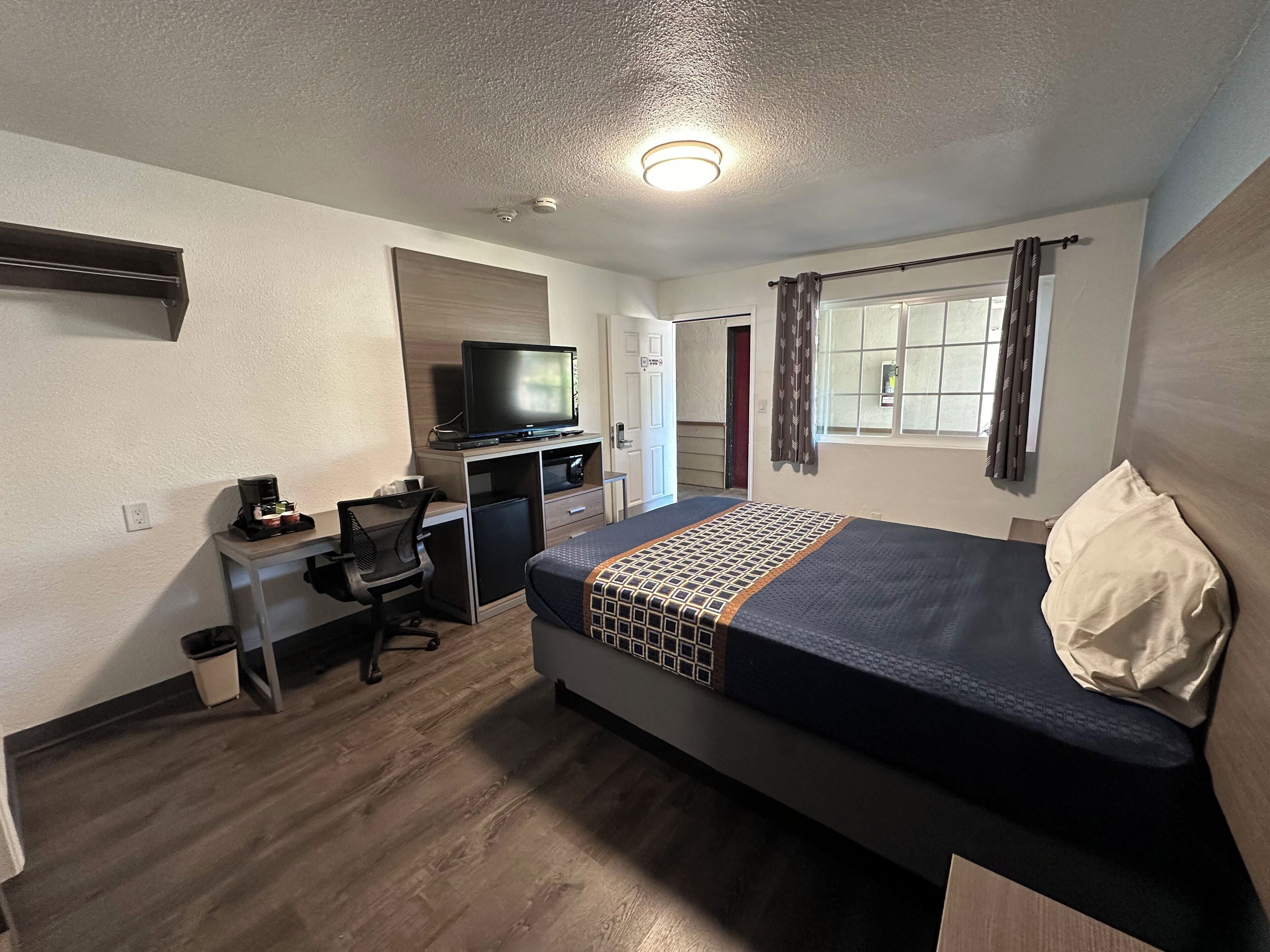A compact hotel room with a single bed, a desk with a computer, a television, and a small refrigerator.