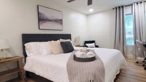 A modern bedroom with a large bed, a ceiling fan, a bedside table with a lamp, a chair, and a window with curtains. - Golf Course Views, Sunsets & Chill Vibes (Lowndes County)