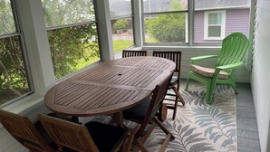 Um alpendre coberto com mesa e cadeiras de madeira, janelas amplas e vista para uma casa e árvores.