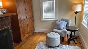 Un salon avec une cheminée, des meubles en bois, un fauteuil gris avec un repose-pieds, une petite table ronde et un lampadaire.