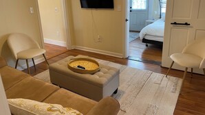 Uma sala de estar com um sofá bege, um banquinho combinando e uma televisão de tela plana na parede.